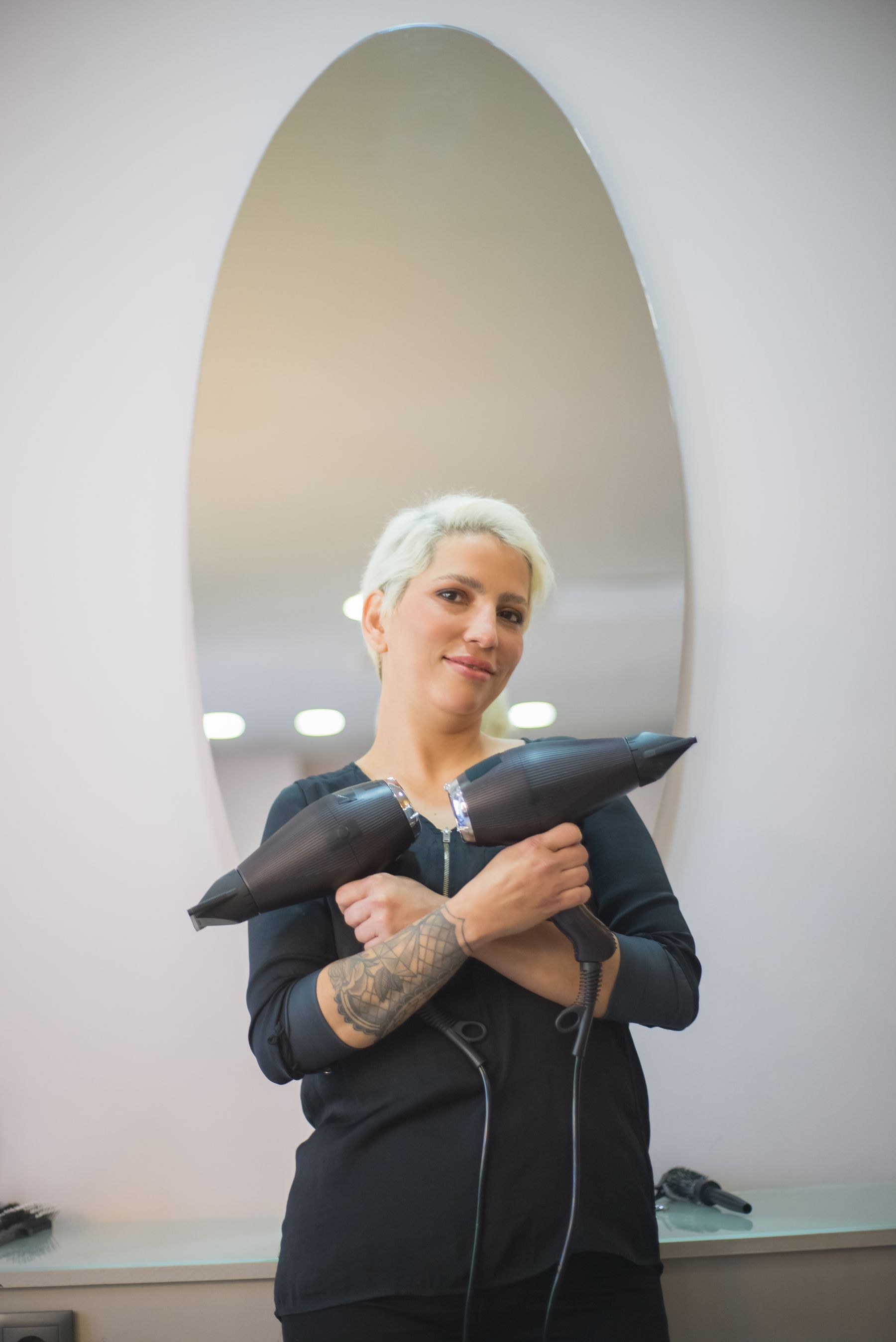 Stylist holding two hairdryers in front of a salon mirror